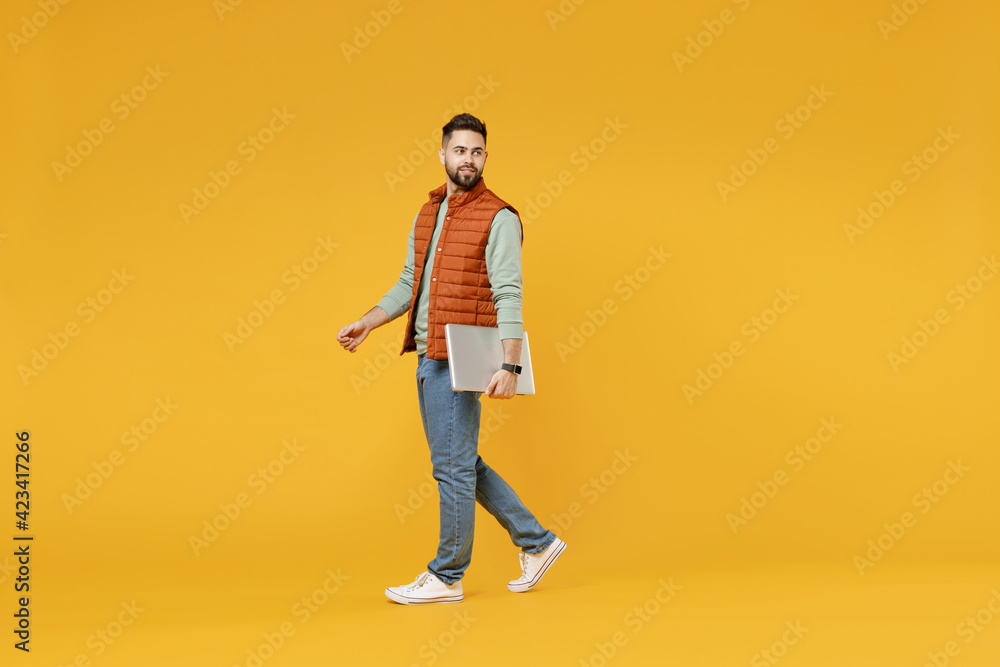 Fototapeta premium Young freelancer happy caucasian fun man 20s wearing orange vest mint sweatshirt glasses using laptop pc computer walking go look aside isolated on yellow background studio. People lifestyle concept.