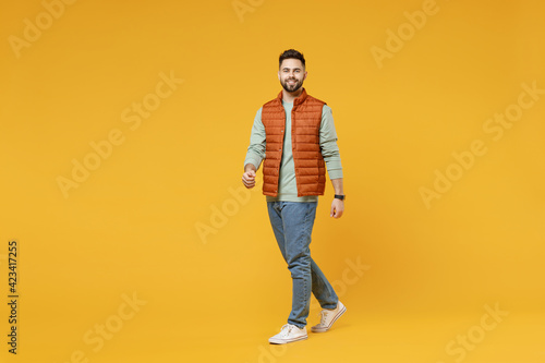 Full length young smiling happy confident smiling cheerful fun caucasian man 20s years old wear orange vest mint sweatshirt walking going looking camera isolated on yellow background studio portrait