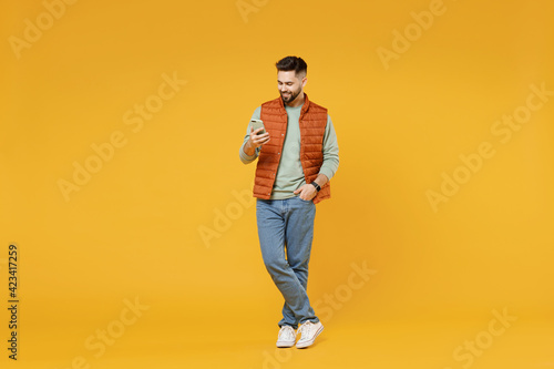 Full length young smiling happy friendly man in orange vest mint sweatshirt using mobile cell phone browsing surfing internet isolated on yellow background studio portrait People lifestyle concept