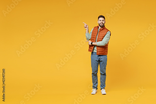 Full length young smiling happy confident smiling cheerful fun caucasian man in orange vest mint sweatshirt point index finger aside on copy space area mock up isolated on yellow background studio