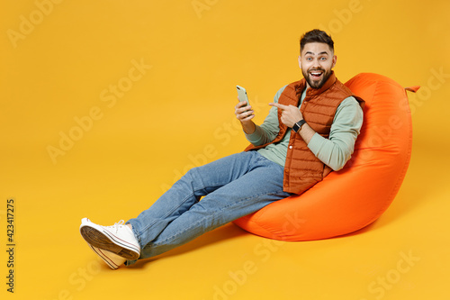 Full length young smiling fun man 20s years old in orange vest mint sweatshirt sitting in beanbag bag chair point index finger on mobile cell phone chat isolated on yellow background studio portrait