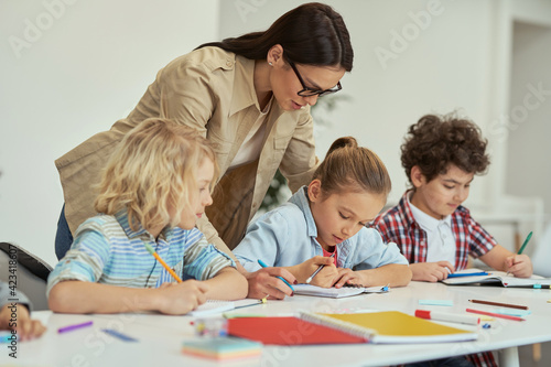 Excellence. Kind young female teacher in glasses helping her little schoolchildren in a classroom. Kids sitting at the table, studying in elementary school