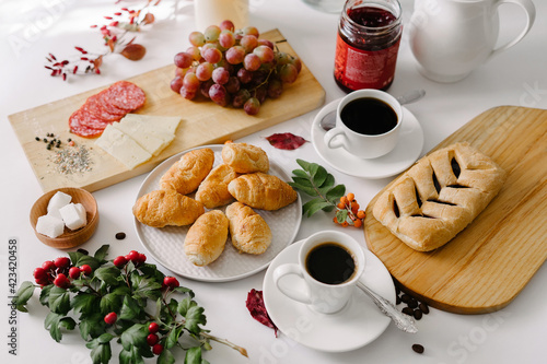 Continental breakfast of mini croissants, pastry, coffee in cups, grapes and jam in a jar, served on white table, decorated with white flowers bouquet