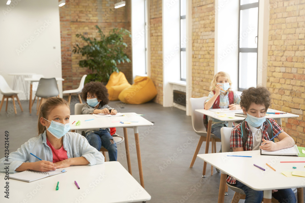 Wear face coverings indoors. School kids wearing protective face mask during coronavirus outbreak studying in elementary school, sitting at the desk in classroom Wear face coverings indoors. School kids wearing protective face mask during coronavirus outbreak studying in elementary school, sitting at the desk in classroom