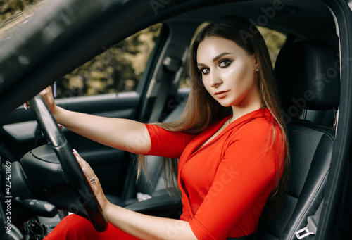 A beautiful young girl in a red overalls sits behind the wheel of a black car on an empty road in the forest © Daria Lukoiko