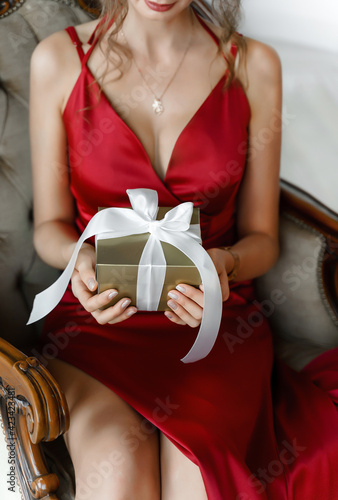 Attractive young woman in a classy red silk dress holds golden gift box with white ribbon