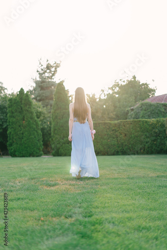 Young beautiful woman with long fair hair in long light blue chiffon dress, walking in the green garden at sunset, view from back
