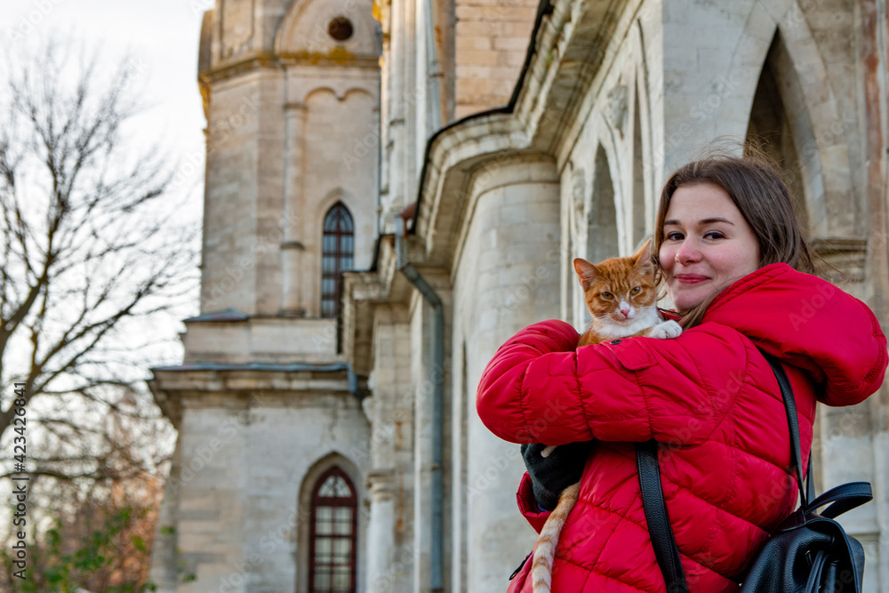 A girl in a red jacket holds a ginger cat in her arms standing next to the wall of an old building