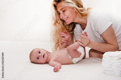 Mother and child on a white bed. Mom and baby boy or girl in diaper playing in sunny bedroom. Parent and little kid relaxing at home. Family having fun together. Bedding and textile for infant nursery