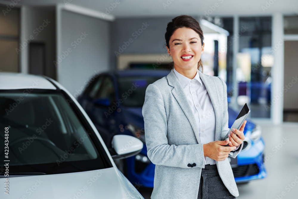 Friendly, smiling female car seller standing in car salon and holding tablet while looking at camera. - obrazy, fototapety, plakaty 