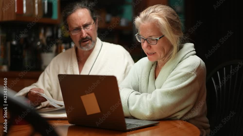 Mature middle age couple doing paperwork using laptop sit at kitchen table in home dining room at night. Mature middle age couple doing paperwork using laptop sit at kitchen table in home dining room at night.