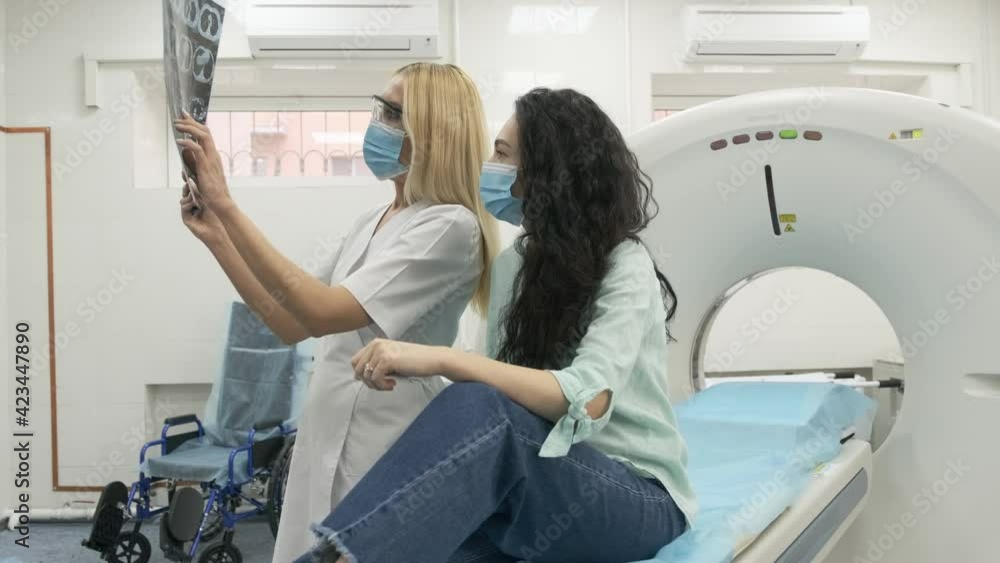 Woman doctor radiologist explains the results of CT scanning for young female patient, showing the snapshot with images, observing and analyzing CT scan in modern clinic beside modern CT scanner Woman doctor radiologist explains the results of CT scanning for young female patient, showing the snapshot with images, observing and analyzing CT scan in modern clinic beside modern CT scanner