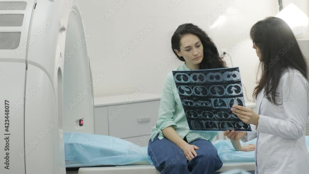 Woman doctor radiologist explains the results of CT scanning for young female patient, showing the snapshot with images, observing and analyzing CT scan in modern clinic beside modern CT scanner Woman doctor radiologist explains the results of CT scanning for young female patient, showing the snapshot with images, observing and analyzing CT scan in modern clinic beside modern CT scanner
