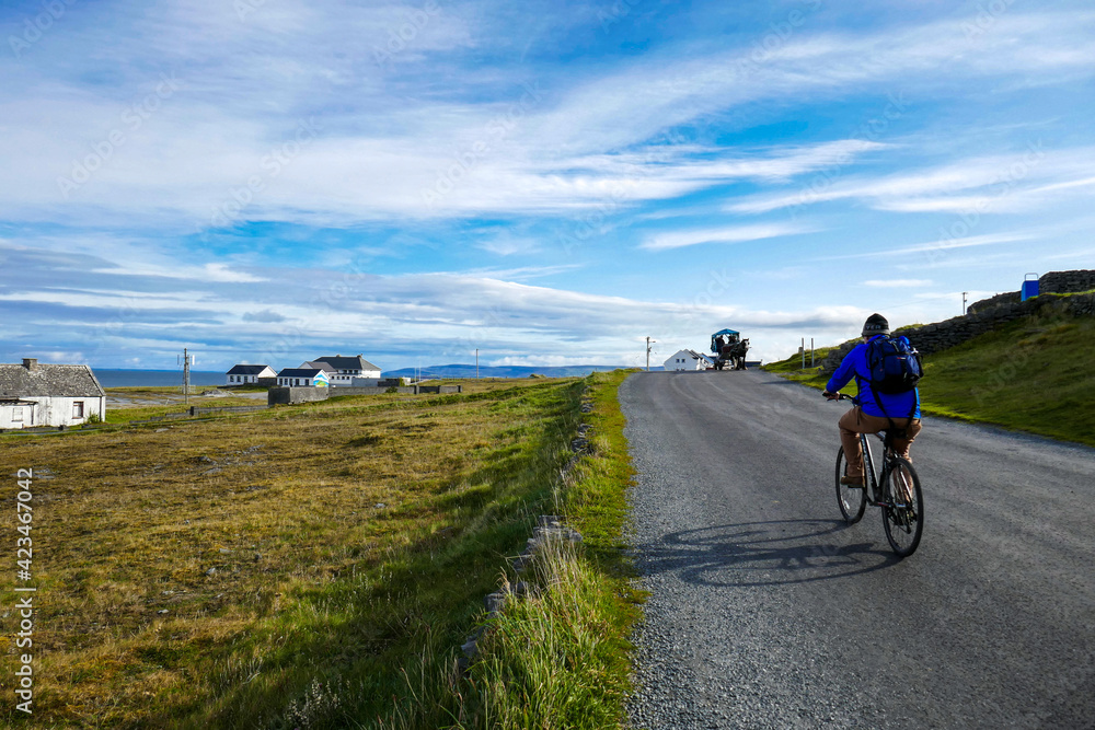 Beautiful Inis Oirr in the Aran Islands. Cyclist on road with cloudy blue sky and field with quaint houses. Beautiful Inis Oirr in the Aran Islands. Cyclist on road with cloudy blue sky and field with quaint houses.