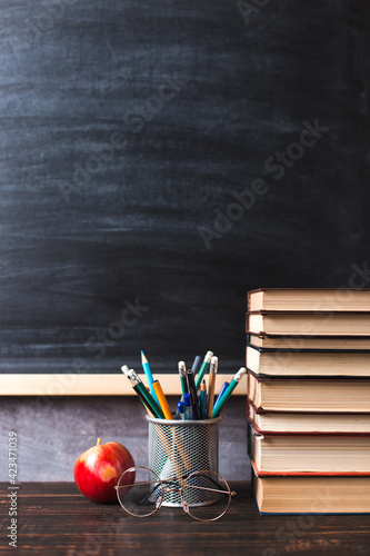 Concept to teacher's day. Pens, apple, pencils, books and glasses on the table, against the background of a chalkboard. Back to school, copy space.