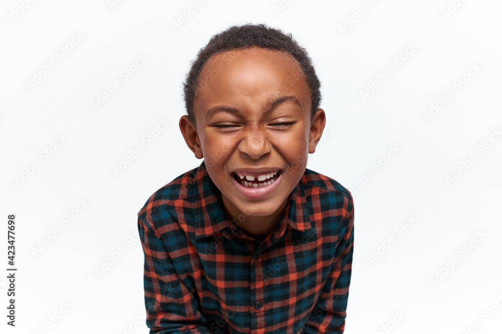 Funny adorable African little boy wearing stylish plaid shirt having cheerful look, closing eyes, laughing out loud, watching favorite cartoon, showing milk teeth. Fun, joy and carefree childhood Funny adorable African little boy wearing stylish plaid shirt having cheerful look, closing eyes, laughing out loud, watching favorite cartoon, showing milk teeth. Fun, joy and carefree childhood