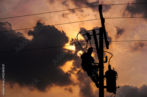 The silhouette of power lineman climbing on an electric pole with a transformer installed. And replacing the damaged hotline clamp, bail clamp, dropout fuse cutout that causes a power outage.