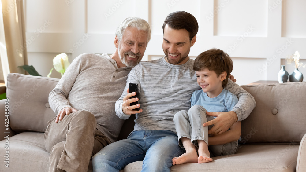 Smiling three generations of men sit rest on sofa in living room talk on video call on smartphone gadget. Happy little Caucasian boy child with father and grandfather use modern cellphone at home.