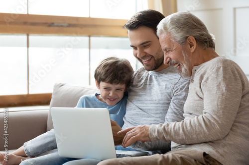 Overjoyed three generations of Caucasian men relax on couch look at laptop screen watch funny video together. Smiling little boy child with young father and grandfather use computer talk on webcam.