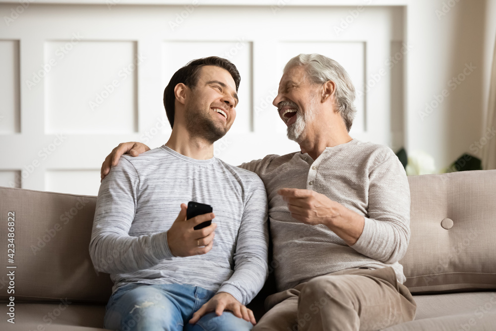 Overjoyed millennial man and senior father relax on sofa at home laugh watching funny video on cellphone together. Smiling old Caucasian dad and adult grownup son have fun using smartphone device.