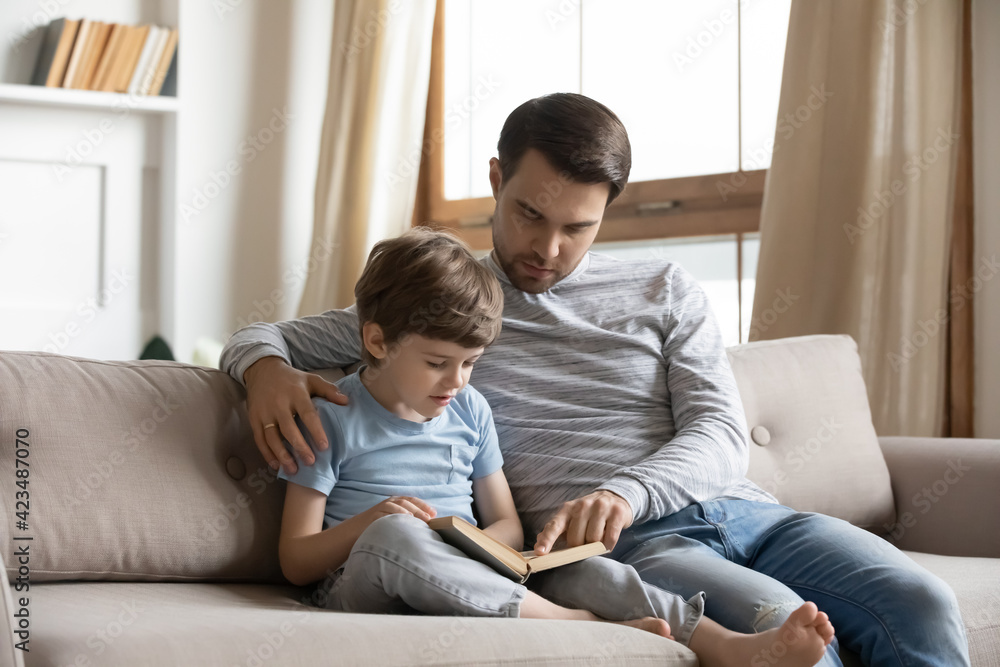 Caring young father sit on sofa with little son teach practice reading book together. Loving Caucasian dad and small 7s boy kid child enjoy textbook story, study at home on weekend. Education concept. Caring young father sit on sofa with little son teach practice reading book together. Loving Caucasian dad and small 7s boy kid child enjoy textbook story, study at home on weekend. Education concept.