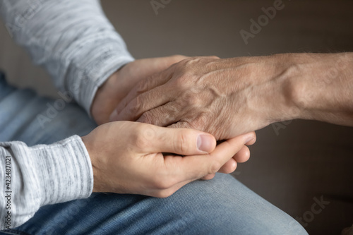 Crop close up of adult grownup son hold elderly father hand show love and care in family relations. Attentive millennial man kid child support comfort unhappy sad mature dad. Bonding, unity concept.