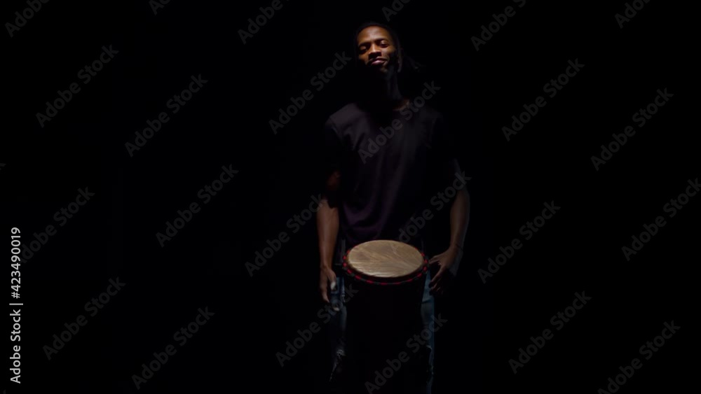 Black man drummer from the darkness smiles and plays on an African drum