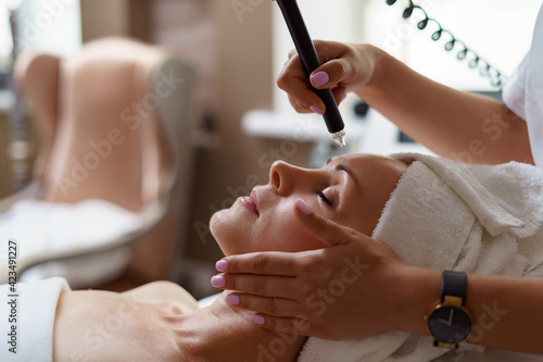 View of doctor cosmetologist doing anti aging procedure in cosmetology office. Satisfied woman in disposable hat lying on couch and relaxing. Working with Apparatus.