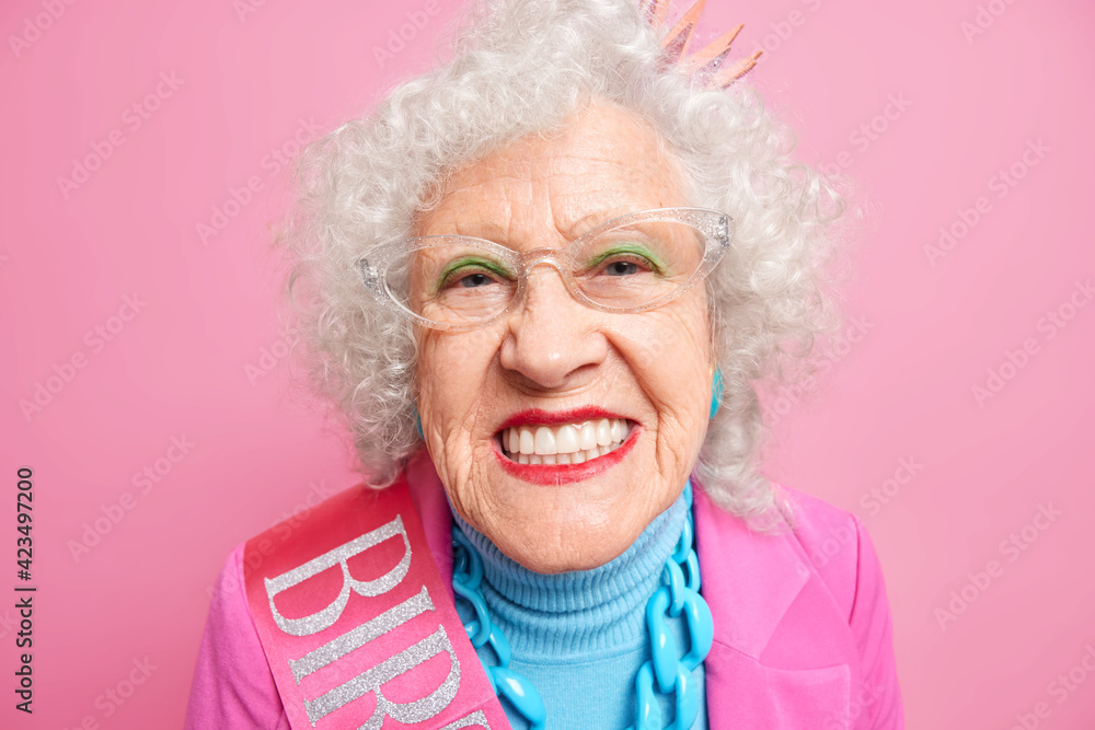 Close up shot of senior grey haired European woman smiles broadly applies bright makeup shows her perfect whit teeth celebrates birthday isolated over pink background. People old age concept Close up shot of senior grey haired European woman smiles broadly applies bright makeup shows her perfect whit teeth celebrates birthday isolated over pink background. People old age concept