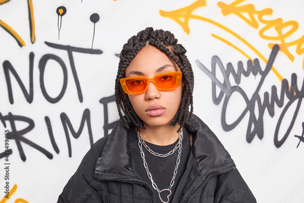Photo of serious teenage hipster girl with dreadlocks wears trendy orange sunglasses and black jacket looks directly at camera has calm expression belongs to hooligan company of friends. Swag - obrazy, fototapety, plakaty 