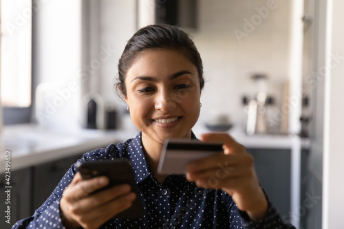 Close up happy Indian woman holding plastic credit card and using phone, shopping, paying online, browsing apps, banking service, smiling satisfied customer making secure internet payment