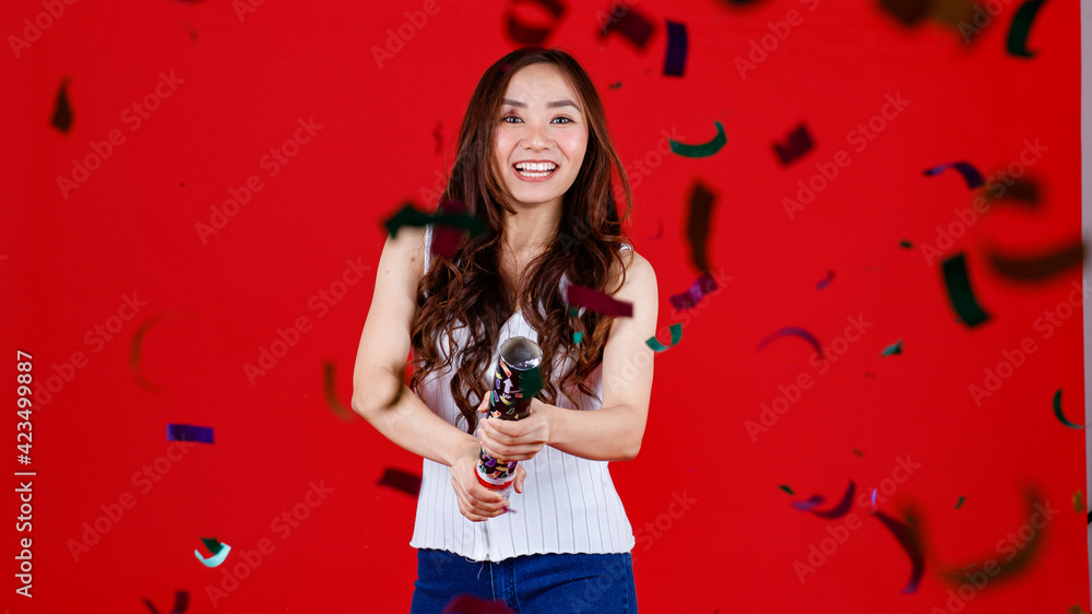 Funny young and cure Asian girl holding and blowing out party poppers with funny and happy. Studio shot on red background Funny young and cure Asian girl holding and blowing out party poppers with funny and happy. Studio shot on red background
