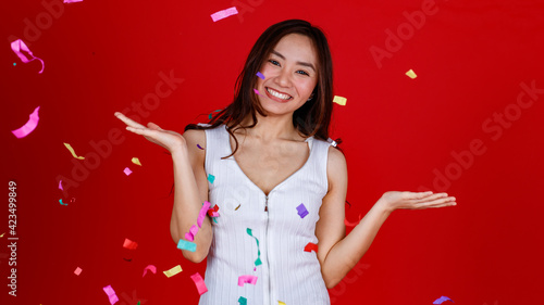 Funny young and cure Asian girl feels excited and cheerful while playing with flying confetti from the above position. Studio shot on a red background