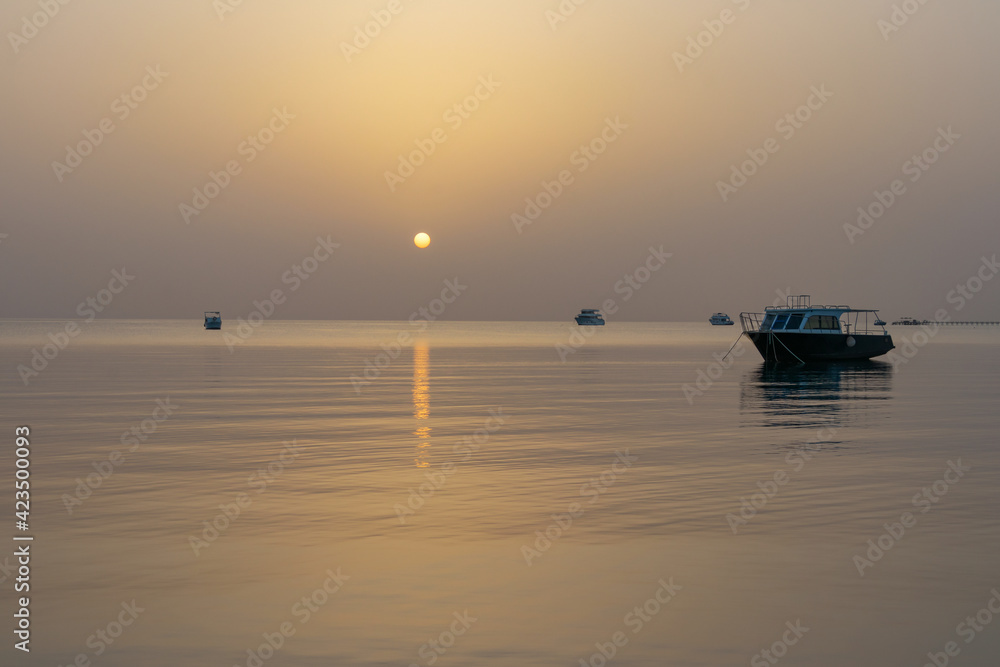 Dawn on the Red Sea in Egypt, Makadi Bay, calm sea, silhouettes of boats, golden disc of the sun on the horizon, reflection of sunlight on the water surface
