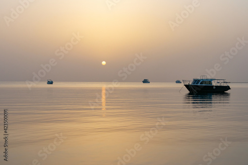 Dawn on the Red Sea in Egypt, Makadi Bay, calm sea, silhouettes of boats, golden disc of the sun on the horizon, reflection of sunlight on the water surface