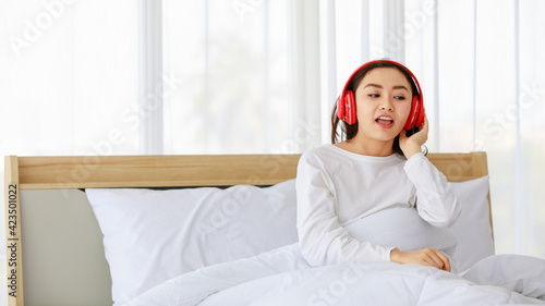 Portrait shot of a young adult Asian woman in casual white sings a song, lying down on the bed and listening to the music with a red headphone. There is copy space at the side of picture
