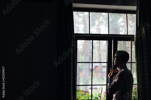 A handsome respectable groom stands at the window in a luxurious hotel room, in a light wedding suit, straightens his shirt.