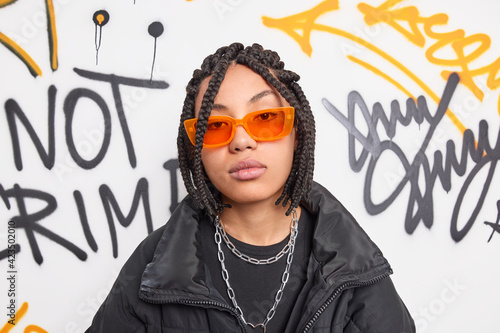 Serious trendy hipster girl wears orange sunglasses black jacket and metal chains around neck belongs to youth subculture poses against graffiti wall. Millennial people style hooliganism concept
