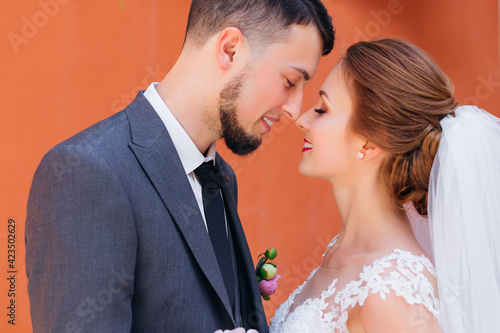 Stylish groom in a gray suit and a beautiful bride in a white dress against the background of an orange wall of an old house gently touch each other.