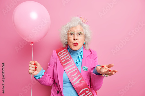 Old people and festive occasion concept. Beautiful wrinkled woman pensioner keeps lips rounded raises palm holds inflated balloon isolated over pink background celebrates birthday enjoys party