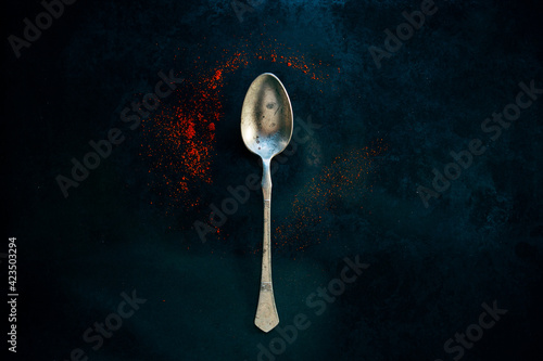 Vintage old teaspoon laying on dark background surrounded with red ground pepper spice, dark and spooky reflection in it