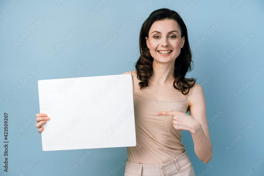 Beautiful girl holding a white sheet of paper points her finger at it on blue background. Space for text, banner, surface for labels, advertising, text. Beautiful girl holding a white sheet of paper points her finger at it on blue background. Space for text, banner, surface for labels, advertising, text.