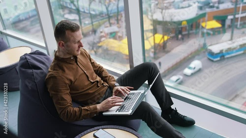 Handsome employee with a laptop in office. Young businessman sitting in a comfortable chair and works near the window with city landscape. Top view. photo