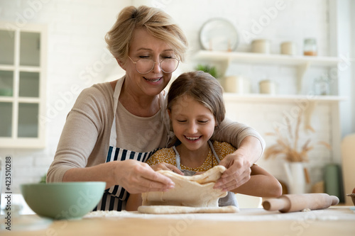 Bilde på lerret Loving mature Caucasian 60s grandmother roll dough bake bread or sweet buns with happy little granddaughter in kitchen