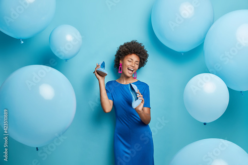 Positive African American woman with curly hair keeps shoes near mouth pretends singing wears dress chooses outfit for party event isolated over blue background with inflated balloons around photo
