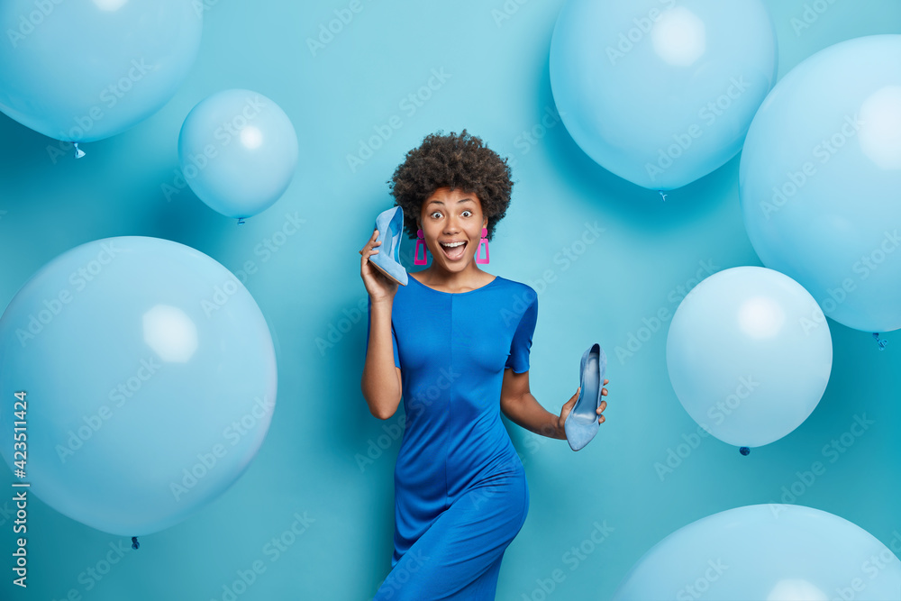 Fototapeta premium Horizontal shot of curly haired young woman wears dress holds high heeled shoes dresses for party poses against blue background with big inflated balloons. People festive event fashion concept