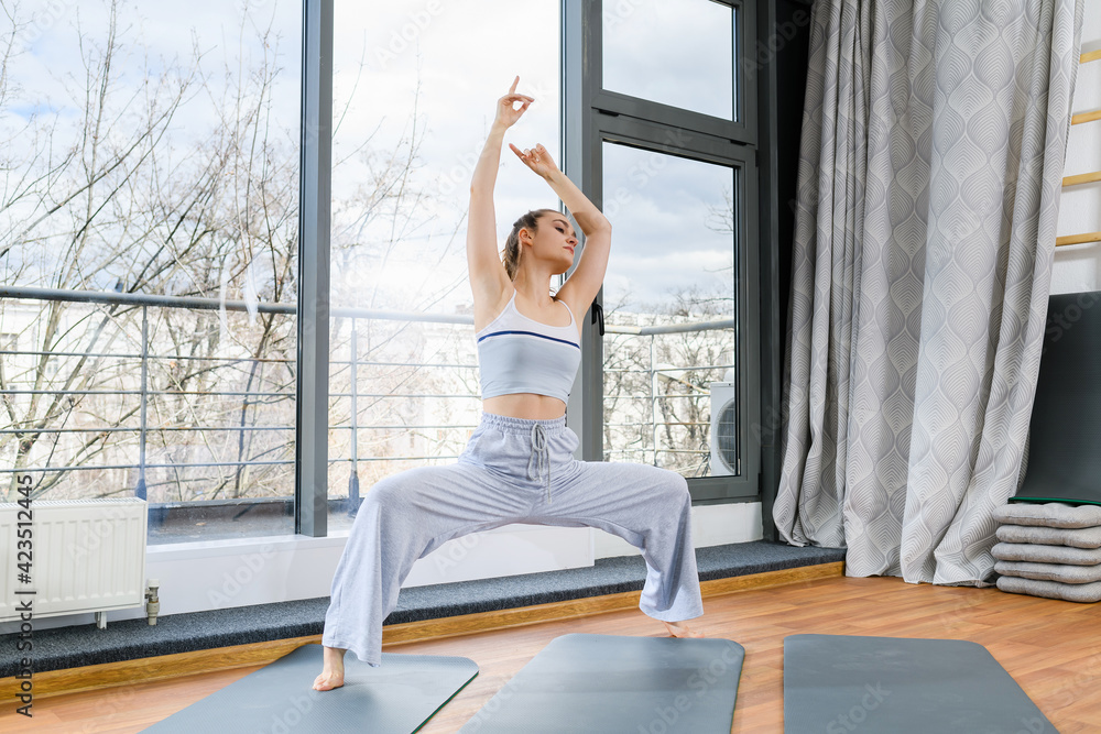 Girl stand in grey sportswear with wide legs on mat and hands up in studio with big window, neutral colors, healthy lifestyle