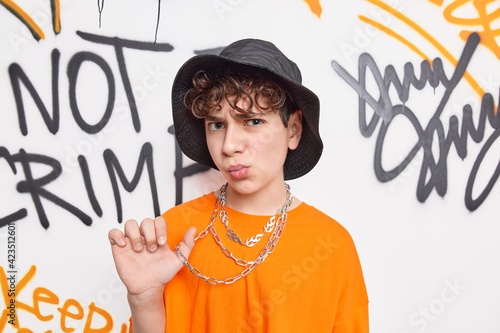 Valokuva Teenage street artist wears hat orange t shirt and chain poses against graffiti wall has discontent expression