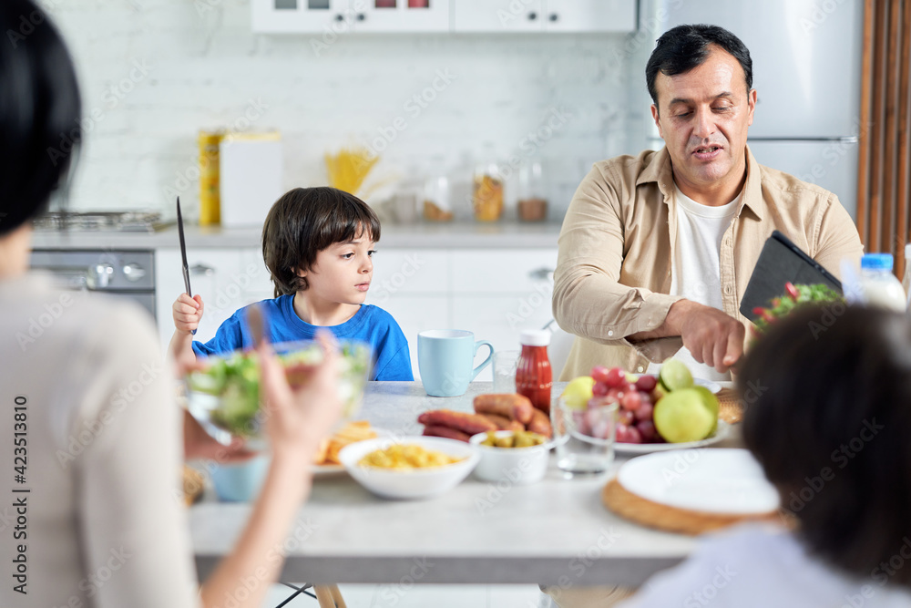 Middle aged father chatting with his child while having breakfast in the morning. Latin family enjoying meal together, sitting at the table in kitchen at home
