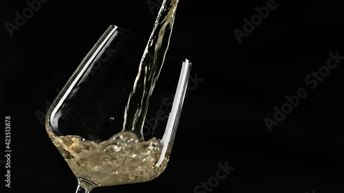 Pouring gold wine into goblet at black background. Close-up of filling wine glass with white wine in super slow motion. White wine forms beautiful wave in glass with copy space photo
